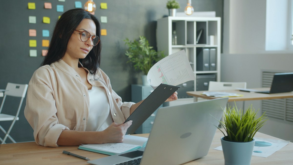 Business owner checking a clipboard of application documents at her laptop before submitting a supplement merchant account file
