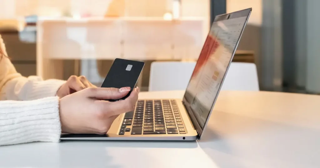 Hands holding a payment card beside a laptop while entering details for a card-not-present transaction online
